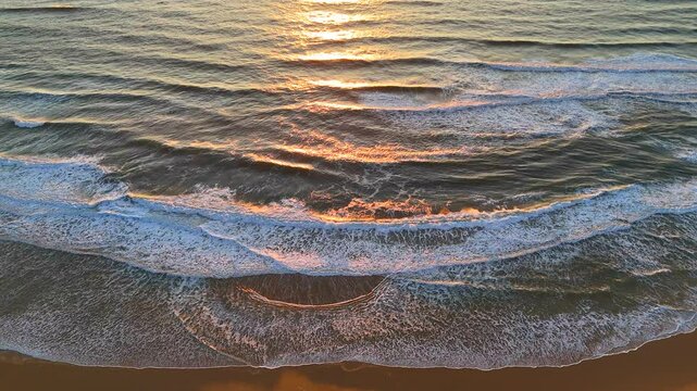Drone captures golden sunset reflecting across textured ocean waves with foamy surf rolling onto a quiet sandy shoreline in soft, peaceful light at the edge of day. 