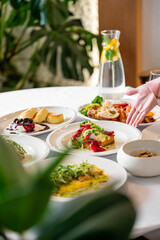 A beautifully arranged table with various fresh dishes, including salads, pastries, and a lemon-infused water pitcher. A hand reaches for a plate, creating a lively and inviting dining scene.
