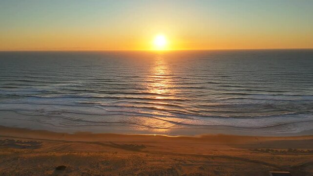 Aerial drone shot of golden sunset over calm ocean, with waves reflecting light and a peaceful sandy beach in the foreground view. A tranquil, scenic coastal moment at dusk.