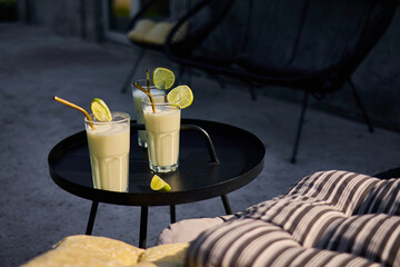 Glasses of lime and condensed milk cocktails sit on a black table in a sunlit terrace. Fresh lime wedges garnish each drink, highlighting a casual summer vibe.