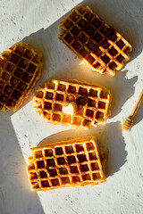 Waffles are arranged on a light surface, with one piece showing a small pat of butter and a honey...