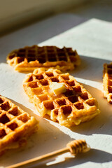 Waffles are arranged on a light surface, with one piece showing a small pat of butter and a honey...