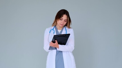 Concentrated Caucasian female young doctor or nurse writing and making notes in notepad on gray background. Concept of medical work.