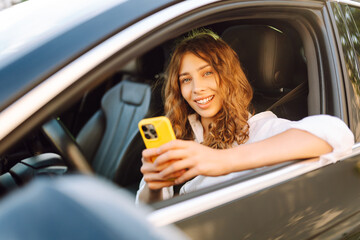 Young woman with curly hair with a phone in her hands driving a car. Happy female driver with a...