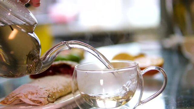 Making hot tea on the kitchen table. Boiling water is poured from the kettle into a cup with a tea bag. Transparent cup of freshly brewed tea on the kitchen table on a sunny day. High quality 4k