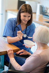 Medical caregiver helping older patient eat soup at home during mealtime