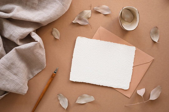 Close-up overhead view of a piece of blank White deckle edge card, envelope and calligraphy pen on a table with a decorative dried leaves and linen fabric on a table