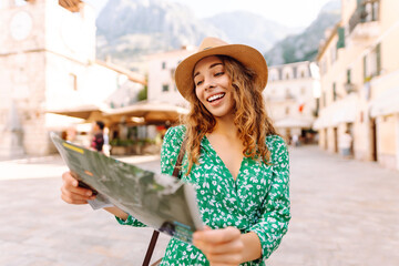 Fototapeta premium Smiling woman in a bright dress looks at a map of the area in the city. Tourist studies navigation of an ancient city against the background of architecture. Travel, vacation concept.