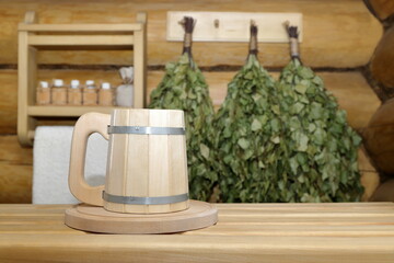 In the sauna interior a wooden beer mug on a tray stands on a plank bench on the background of a log wall with dry birch brooms and shelf with bottles of aromatic oils and white towel.