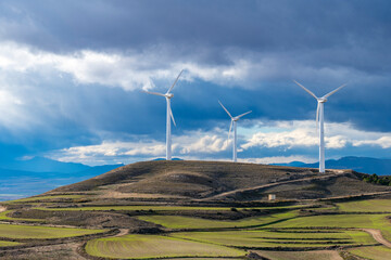 Wind turbines on hilltops with stormy clouds in Zaragoza, vibrant green fields create contrast with environmental technology on the horizon