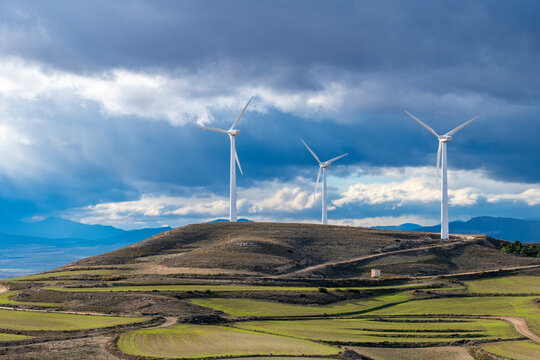 Wind turbines on hilltops with stormy clouds in Zaragoza, vibrant green fields create contrast with environmental technology on the horizon