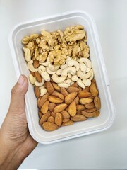 A capture of hand holding a white box of dryfruits containing halved walnuts, almonds and cashews. White backdrop enhancing the focus on isolated box of dryfruits.