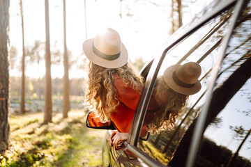 Portrait of a young woman with curly hair enjoying the sunset from the car window. Happy traveler feeling freedom from the car. Freedom, adventure concept.