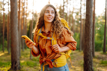 Cute female traveler with a hiking backpack and a phone in her hands walks through a sunny forest....