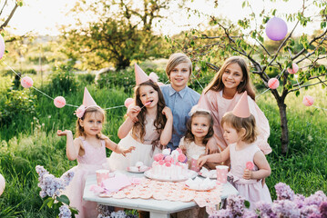 Summer outdoor kids birthday party. Group of happy Children celebrating birthday in park. Children blow candles on birthday cake. Kids party pink pastel decoration and food. Presents and sweets.
