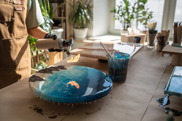 Unrecognizable woman artist use gas torch to remove bubbles from round epoxy resin artwork in...