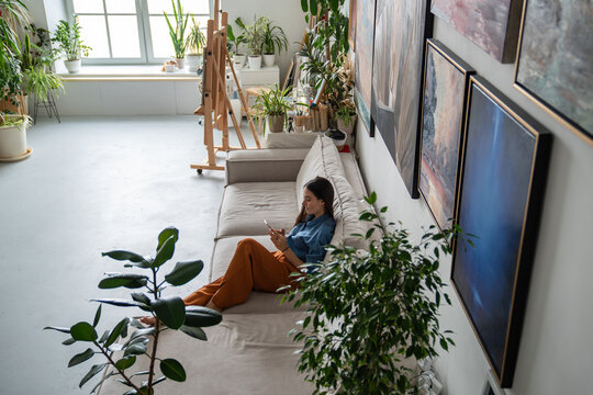 Calm woman artist using smartphone on sofa, pauses during work and reviewing schedule for art school and studio activity. Planning class work, upcoming creative tasks, social media blog, surfing net