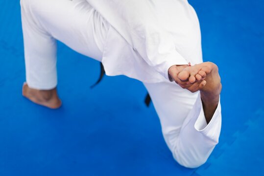 A martial artist in a white gi is captured from above on a blue mat pulling one foot backward while kneeling in a deep stretch during a martial arts flexibility routine indoors