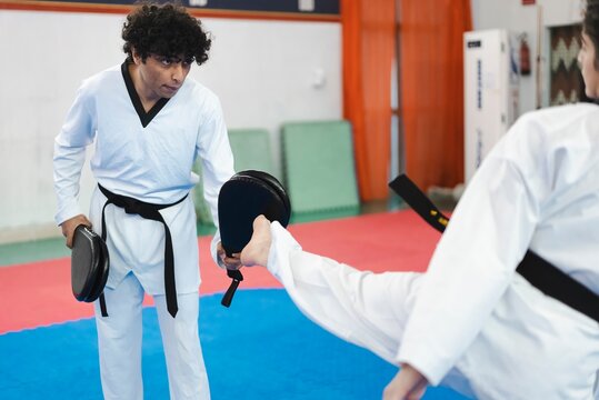 A martial artist in a white gi delivers a forward kick to a black target pad held by a trainer while both wear black belts and train on blue and red mats in a well-lit dojo room
