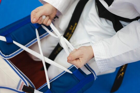 A martial artist wearing a black belt adjusts and ties white laces on a blue padded chest protector while seated on a blue mat during taekwondo training indoors