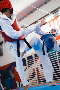 A female martial artist in white gear and blue chest protector lands a kick on the red chest guard of her male opponent during taekwondo sparring on a blue and red mat