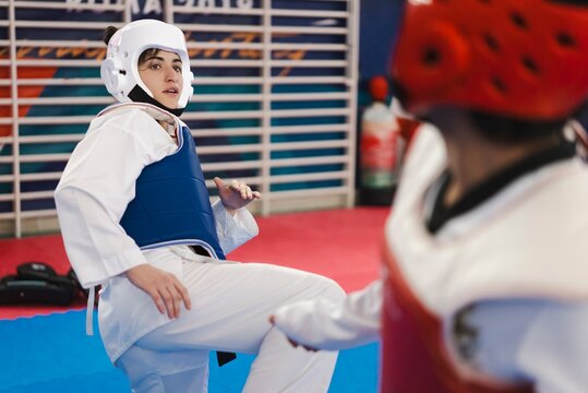 A female martial artist wearing a blue chest protector and white helmet delivers a high kick to her opponent in red gear during a taekwondo sparring session on red mats