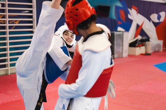 A female martial artist wearing a blue chest protector and white helmet delivers a high kick to her opponent in red gear during a taekwondo sparring session on red mats