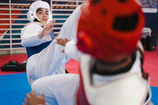 A female martial artist wearing a blue chest protector and white helmet delivers a high kick to her opponent in red gear during a taekwondo sparring session on red mats