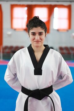 A female martial artist in a white uniform with a black belt stands confidently on a blue mat in a well-lit training room with orange curtains and red chairs in the background