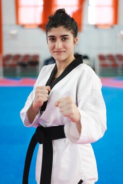 A young female martial artist in a white gi and black belt raises her fists in a focused fighting stance while standing on a blue mat inside a brightly lit training room