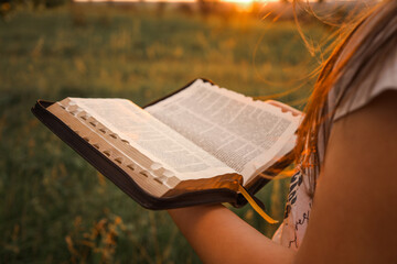 Close-up of a girl reading the Bible outdoors during golden hour. The soft sunset light gently...
