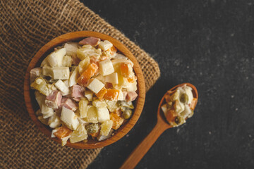 Top view of Olivier salad served in a wooden bowl on a cutting board with a wooden spoon, on a black rustic background. Traditional Slavic dish made with potatoes, ham, peas, and eggs