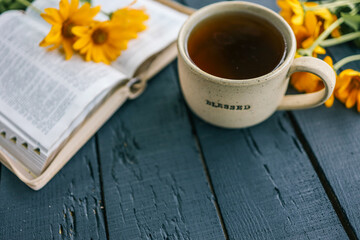 Cup of tea with “BLESSED” inscription next to an open Bible and yellow flowers on a rustic dark...