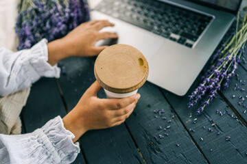 Child working on a laptop while holding a cup with a wooden lid. Lavender flowers lie on a rustic dark wooden table, creating a cozy and peaceful workspace
