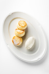 Three golden-brown cheese pancakes on a white plate, accompanied by a scoop of creamy white cheese, creating an appetizing and inviting presentation.