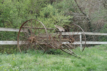 Old rusted agricultural machine with iron wheels overgrown by spring grass and wildflowers in rural Serbia. Abandoned vintage farm equipment near wooden fence in natural setting.