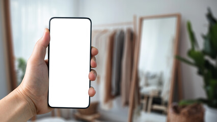 Hand Holding Blank Smartphone Display in Modern Room with Mirror and Stylish Clothes Rack in Soft Natural Light. Agnostic