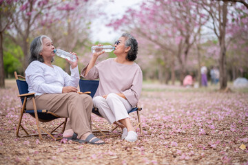 Two elderly women are sitting outdoors chairs in a park-like setting with pink scattered on the...