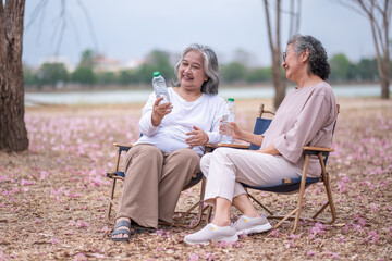 Two elderly women are sitting outdoors chairs in a park-like setting with pink scattered on the...