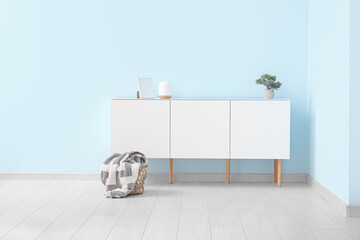Modern chest of drawers with houseplant, blank picture and basket near blue wall in room