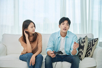 Obraz premium A young Asian man and woman are sitting on a living room sofa, holding game controllers, watching TV, and happily playing a game