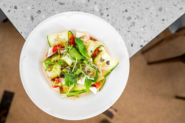 A fresh salad featuring sliced cucumbers, cherry tomatoes, and greens, drizzled with dressing, served in a modern white bowl on a stylish table