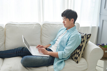 Obraz premium A young Asian man wearing a blue shirt and jeans is sitting on the living room sofa, working on a laptop.