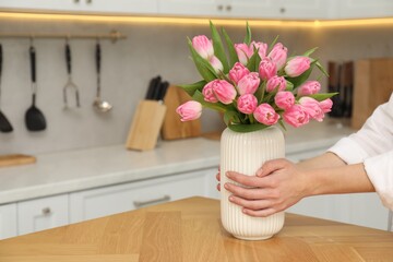 Woman holding vase with bouquet of beautiful tulips at wooden table in kitchen, closeup. Space for...