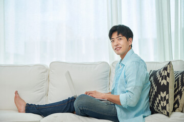 A young Asian man wearing a blue shirt and jeans is sitting on the living room sofa, working on a laptop.