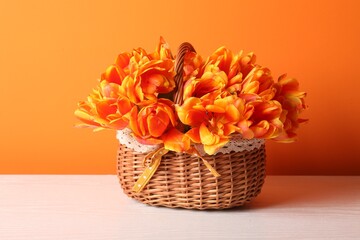 Beautiful tulips in wicker basket on white wooden table against orange background