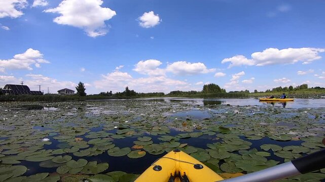 Kayaking along river through growing white lily nymph on sunny summer day. Person floating in boat on river surface of water reflecting white clouds on calm day. Tourism. Active lifestyle. POV