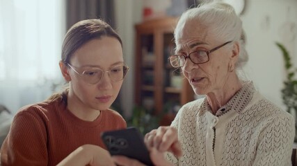 Young woman teaching elderly grandmother how to use smartphone, helping her to explore modern technology at home