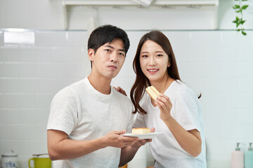 Fototapeta premium A lovely couple in white T-shirts sits the woman on the kitchen table, sharing a plate of castella cake.