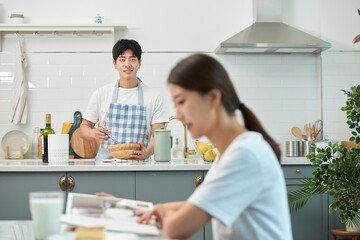 The young husband is cooking in the kitchen while his wife sits at the dining table reading a book